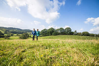 Zwei Personen wandern durch den Naturpark Hüttener Berge. Sie stehen auf einer Wiese. Im Hintergrund sieht man ein paar Bäume und die Landschaft.