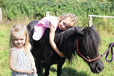 Ein kleines Mädchen liegt auf dem Rücken eines schwarzen Pferdes. Ein anderes Mädchen steht daneben und schaut in die Kamera.