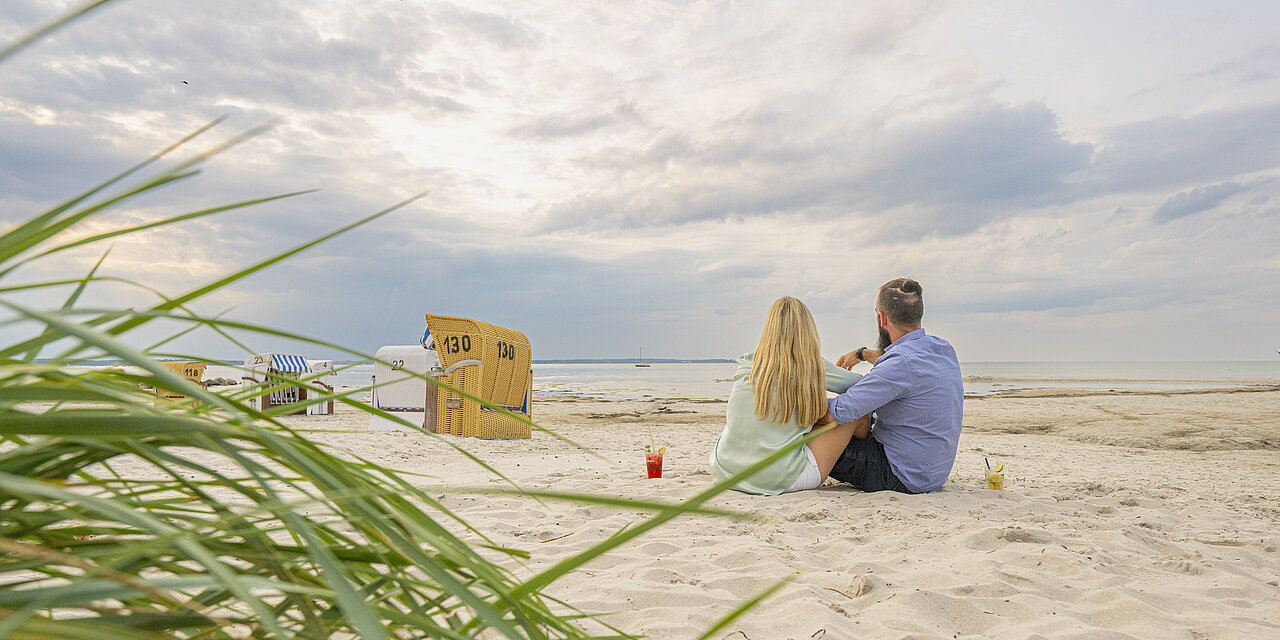 Ein Mann und eine Frau sitzen dicht beieinander im Sand, vor ihnen das Meer. 