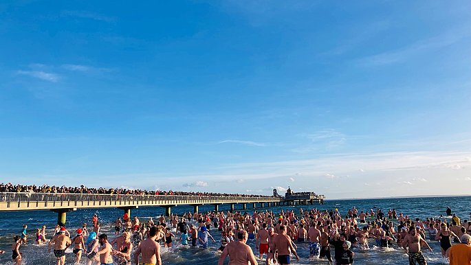 Viele Menschen laufen im Badeanzug bekleidet in die Ostsee. Rechts ist die Seebrücke von Grömitz zu sehen.