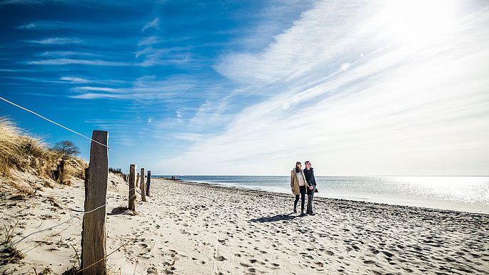 Ein Pärchen spaziert über den Sanstrand entlang der Ostseeküste. Die Sonne scheint und der Himmel ist blau.