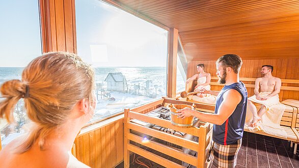 Der Saunameister schüttet Wasser aus einem Sauna-Topf aus Holz auf den Saunaofen. Im Hintergrund sieht man die Saunagäste. Aus dem Fenster kann man die Ostsee erkennen.