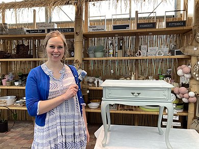 Eine Frau mit blauer Strickjacke und Pinsel in der Hand steht in ihrem Geschäft neben einem hell gestrichenen Nachttischchen, das sie anpinselt.