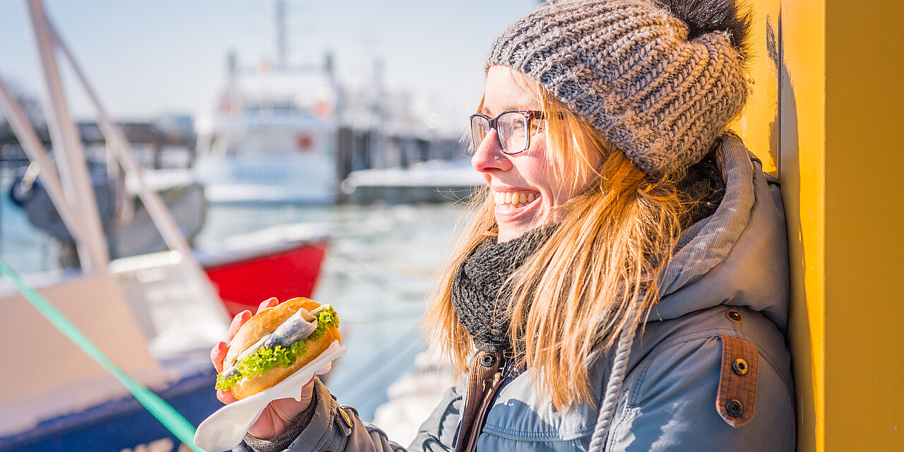 Frau am Hafen an der Ostsee lächelt und hält ein Fischbrötchen in der Hand