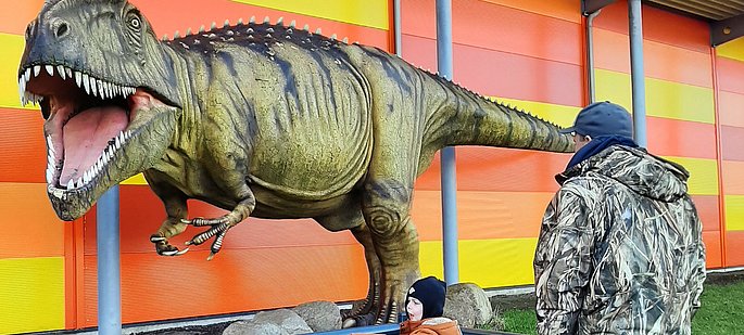 Eine große Dino-Statur steht vor der Galileo Wissenswelt. Ein Papa mit seinen Kindern schaut sich den Dino an.