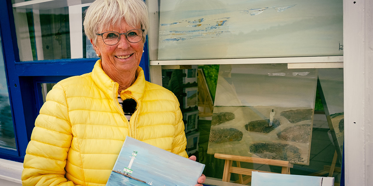 Eine Frau in gelber Steppjacke hält ein Bild in den Händen. Hinter ihr ein weißgestrichener Kiosk am Heikendorfer Strand.