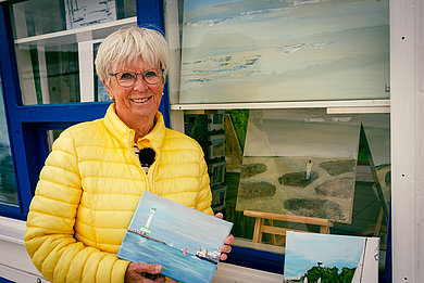 Eine Frau in gelber Steppjacke hält ein Bild in den Händen. Hinter ihr ein weißgestrichener Kiosk am Heikendorfer Strand.