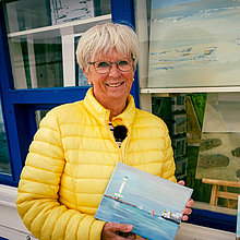 Eine Frau in gelber Steppjacke hält ein Bild in den Händen. Hinter ihr ein weißgestrichener Kiosk am Heikendorfer Strand.