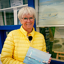 Eine Frau in gelber Steppjacke hält ein Bild in den Händen. Hinter ihr ein weißgestrichener Kiosk am Heikendorfer Strand.