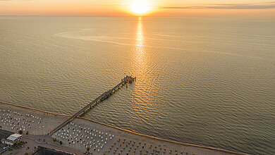 Luftbildannahme von der Landschaft von Dahme im Sonnenaufgang. Im Linken Bildrand ist der Strand mit den vielen weißen Strandkörben zu sehen. Die Seebrücke reicht auf die ruhige Ostsee hinaus. Am oberen Bildrand ist der Horizont mit der aufgehen Sonnenaufgang zu sehen.