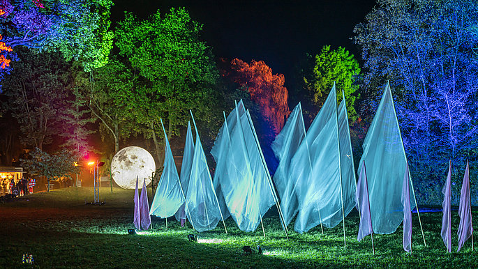 Die Bäume im Kurpark werden mit verschiedenen Lichtern angestrahlt. Manche Bäume im Hintergrund leuchten Blau, andere Grün, oder Rot. Vor den Bäumen in der Mitte sind riesengroße (fast so große wie die Bäume) Fahnen zu sehen. Die Fahnen leuchten in einem durchsichtigem Türkise.