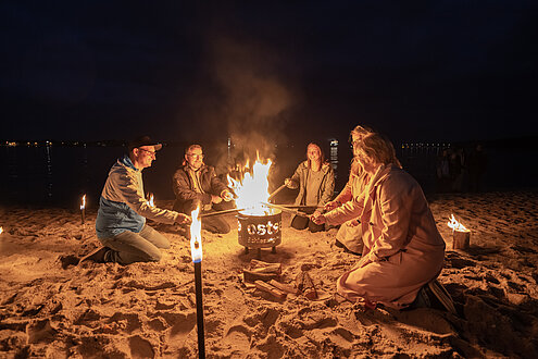 Eine Gruppe von Menschen sitzt um den Feuertopf (Lagerfeuer) herum und wärmt sich am dem Feuer.