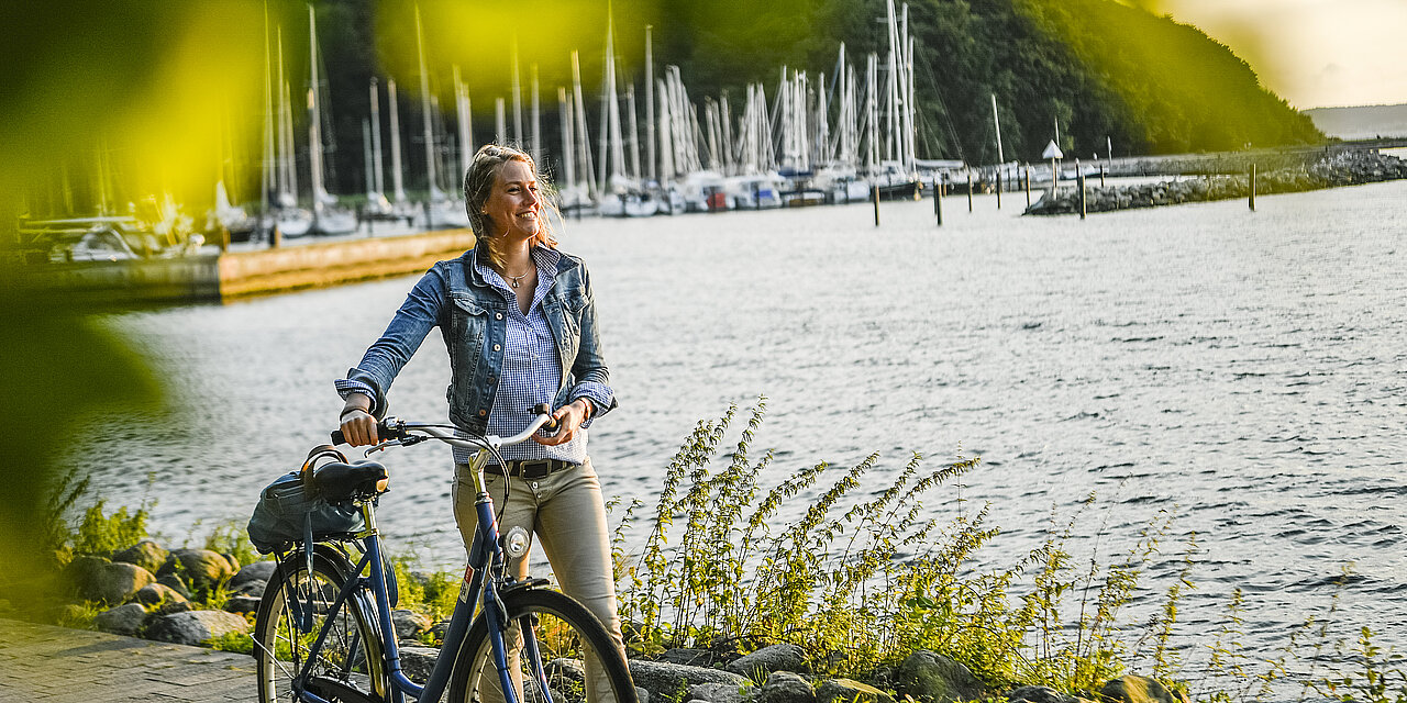Frau schiebt ihr Fahrrad am Hafen entlang. Im Hintergrund sieht man viele Segelboote