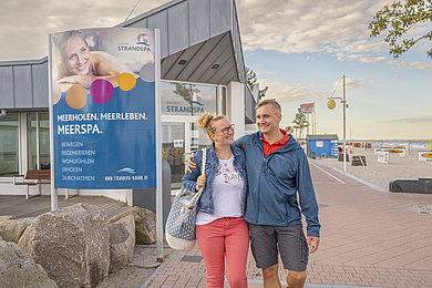 Paar spaziert am Strand in Dahme. Neben ihnen ein Gebäude und davor ein großes Schild vom Strandspa.