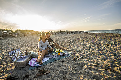Ein Pärchen sitzt am Strand auf einer Picknickdecke und schaut auf die Ostsee. Im Hintergrund geht die Sonne unter.
