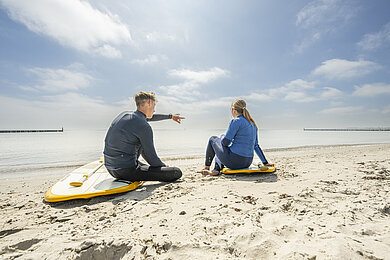 Ein Mann und eine Frau sitzen auf ihren SUP Boards am Strand und schauen auf die Ostsee