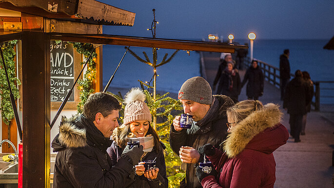 Vier Freunde stehen an einer Glühweinbude und trinken warmen Glühwein. Im Hintergrund ist die Ostsee zu sehen. 