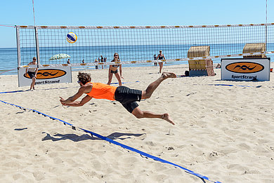 Volleyballspieler auf dem Sportstrand in Dahme spielen Volleyball. Ein Mann springt einem BAll hinterher. Im Hintergrund das Meer.