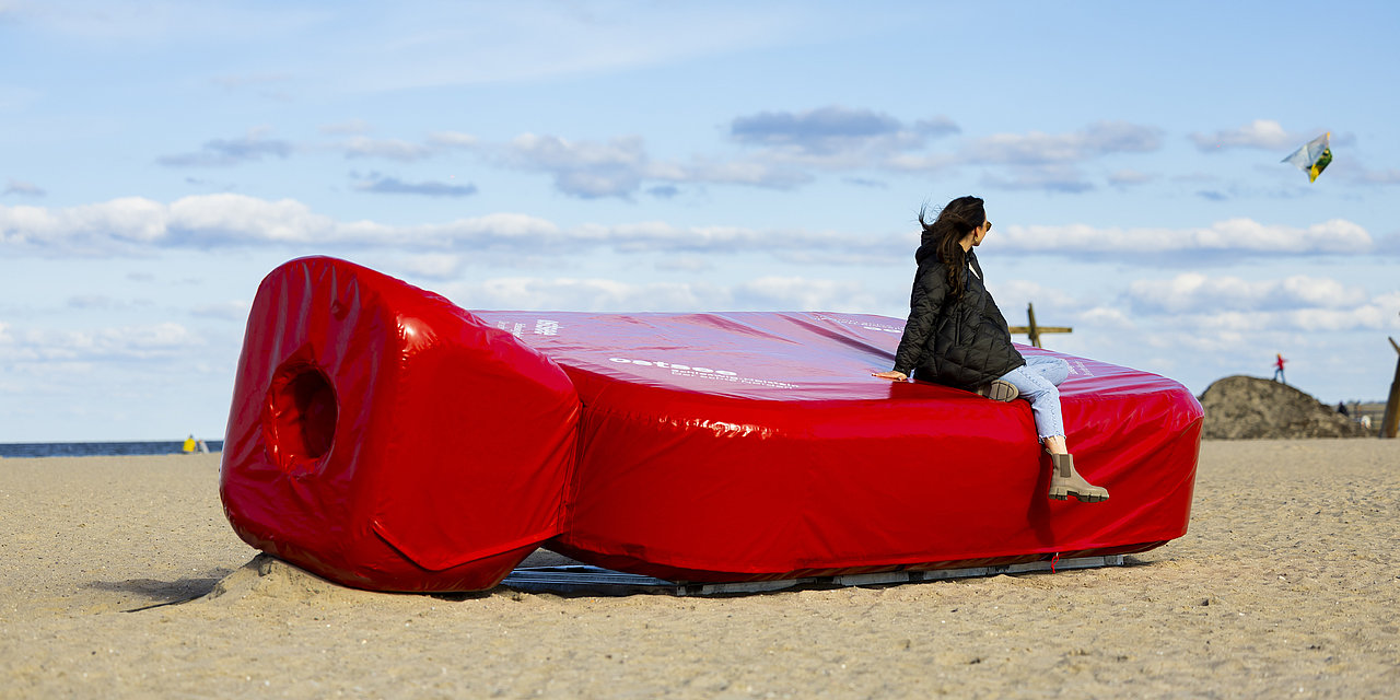 Frau sitzt auf der Riesenwärmflsche Wärmy am Ostsee Strand. Der Wind weht durchs Haar.