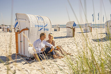 Paar sitzt hinter einem Strandkorb mit den Rücken an der Rückseite des Strandkorbes gelehnt am Strand. Sie genießen ihr Frühstück an einem Sommermorgen. Bei halten ein Croissant und eine Tasse Kaffee in der Hand.
