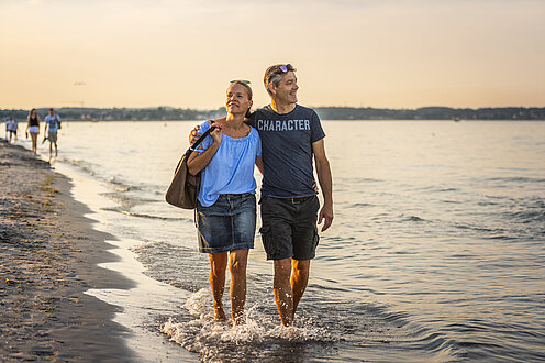 Ein Mann und eine Frau gehen am Strand spazieren. Sie sind barfuß und gehen im flachen Wasser, er hat den Arm um ihre Schultern gelegt,