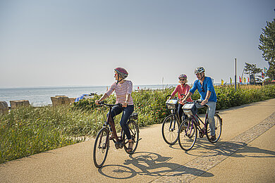 Drei Radfahrer fahren die Promenade in Kellenhusen entlang. Im Hintergrund sieht man den Strand mit seinen Strandkörben und die Ostsee.
