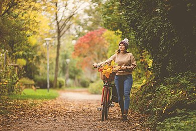 Frau schiebt ihr Fahrrad durch einen Wald. Die Blätter leuchten in herbstlichen Farben.