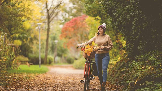 Frau schiebt ihr Fahrrad durch einen Wald. Die Blätter leuchten in herbstlichen Farben.
