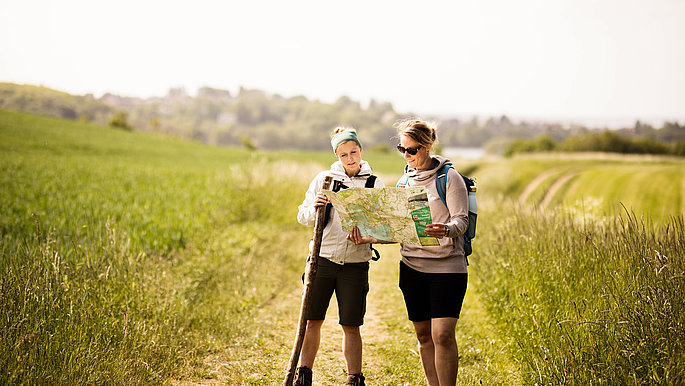 Zwei Frauen wandern auf einem Wanderweg zwischen Feldern und Wiesen. Sie bleiben gerade stehen, um auf eine Wanderkarte zu schauen.
