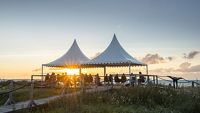 Viele Menschen sitzen auf der Terrasse, welche mit 2 weißen Pavillons ausgestattet sind und schauen den Sonnenuntergang über der Ostsee an. Ein Kiter fährt auf dem Wasser. 
