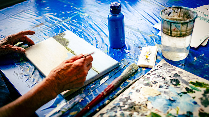 Eine Nahaufnahme. Auf dem Ausschnitt ist ein Bild, das auf einem blauen Tisch liegt. Eine Hand mit einem Pinsel, die gerade auf das Bild malt.