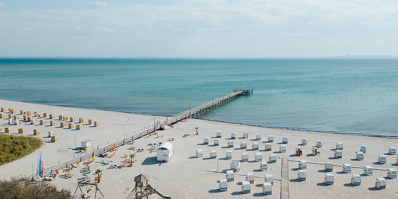 Zu sehen ist der weiße Sandstrand von Pelzerhaken mit Strandkörben und einem Steg ins Wasser.