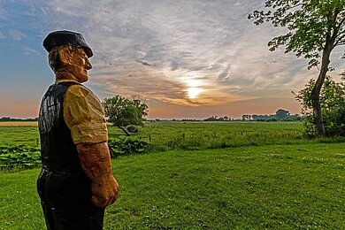 Eine Figur, die einen Bauern darstellt, schräg von der Seite und von hinten. Im Hintergrund leuchtet die Sonne und geht unter.