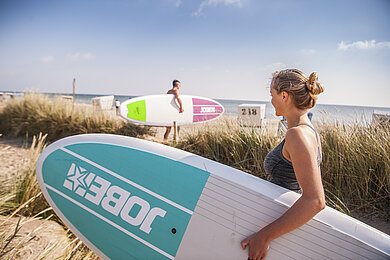 Eine Frau mit einem SUP Board unter dem Arm geht zum Wasser am OStseestrand