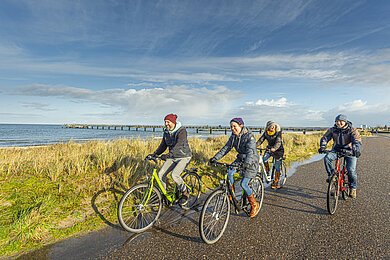 Eine Gruppe Radfahrer fahren eine Straße entlang, links im Bild eine wiese, dahinter das Meer.
