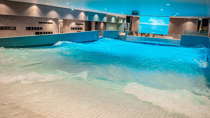 Zu sehen ist das Wellenbad. Am Ende des Becken ist eine Leinwand aufgebaut. Der Beamer der unter der Decke hängt, projiziert eine Strandlandschaft auf die Leinwand 
