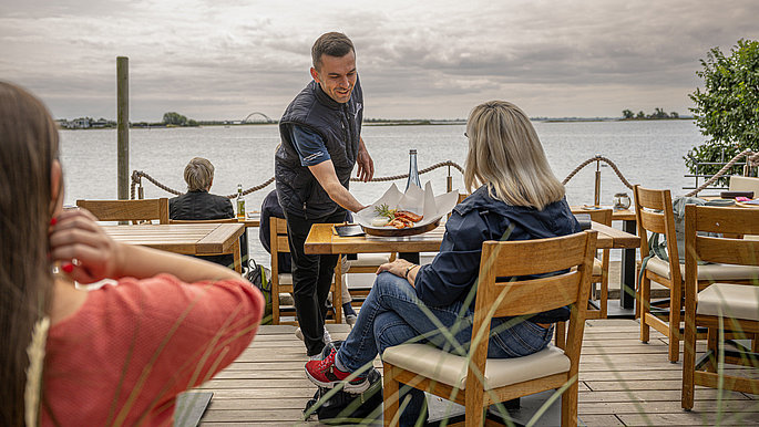 eine Frau sitzt am Tisch in einem Restaurant draußen und schaut aufs Meer. 