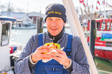 Ein Fischer mit Fischbrötchen in der Hand steht am Hafen