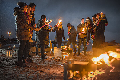 Viele Menschen stehen mit einer Fackel um den Feuerkorb am Strand.