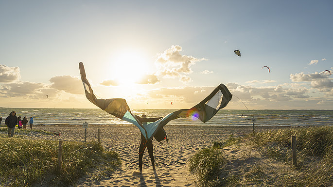 Am Strand steht eine Person mit einem orangen Kite, den er in die Luft hält.