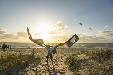 Am Strand steht eine Person mit einem orangen Kite, den er in die Luft hält.