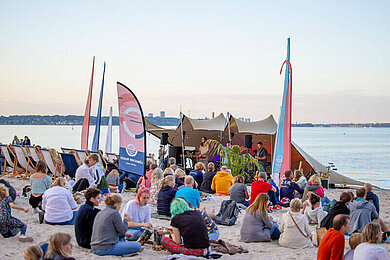 Menschen sitzen am Strand, vor dem Meer eine Bühne auf der Musik gespielt wird. 