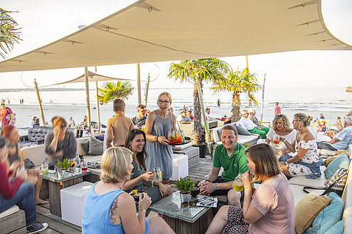 Eine gut besuchte Strandbar in Scharbeutz. Viele Menschen sitzen in Liegestühlen, die Füße im Sand. Ein heller Sonnenschirm ist aufgespannt. 