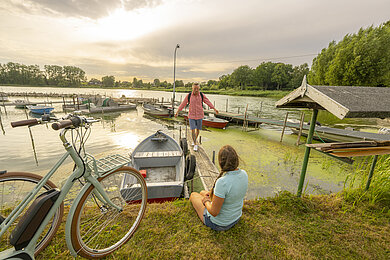 Paar macht während der Radtour eine Pause am Passader See. Dort liegen viele kleine Fischerboote. Die Frau sitzt an der Hafenkante, der Mann balanciert auf dem Steg zwischen den Booten.