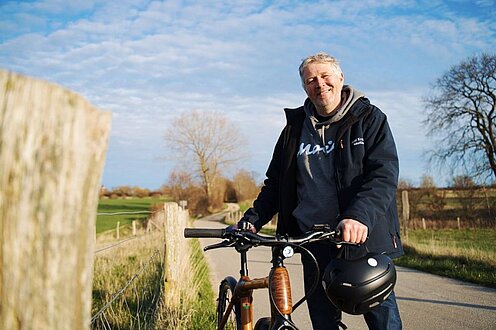 Axel Kramer steht mit seinem Fahrrad auf einer geteerten Straße. Links und rechts sind Felder und Wiesen.