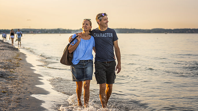 Paar spaziert bei Sonnenuntergang entlang der Ostseeküste. Sie schauen auf das ruhige Wasser.