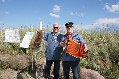 Zwei Männer in blau-weiß-gestreiften Hemden, einer mit Seemannsmütze und Akkordeon, sie stehen vor den Dünen.