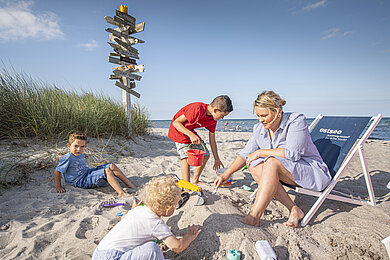 Familie, Mutter mit drei Kindern, sitzt am Strand. Die Mutter sitzt auf einen Beachstuhl, die Kinder spielen vor ihr im Sand mit Sandspielzeug.