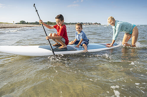 Eine Mutter mit ihren Kindern fährt SUP im Uferbereich der Ostsee. Sie steht nur bis zu den Knien im Wasser und schiebt das SUP von hinten an. Die Kinder sitzen auf dem SUP. Das ältere Kind sitzt vorne und versucht mit dem Paddel mitzuhelfen.
