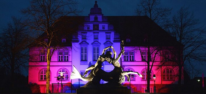 Ein weißes Giebelgebäude erstrahlt in lila Licht. 